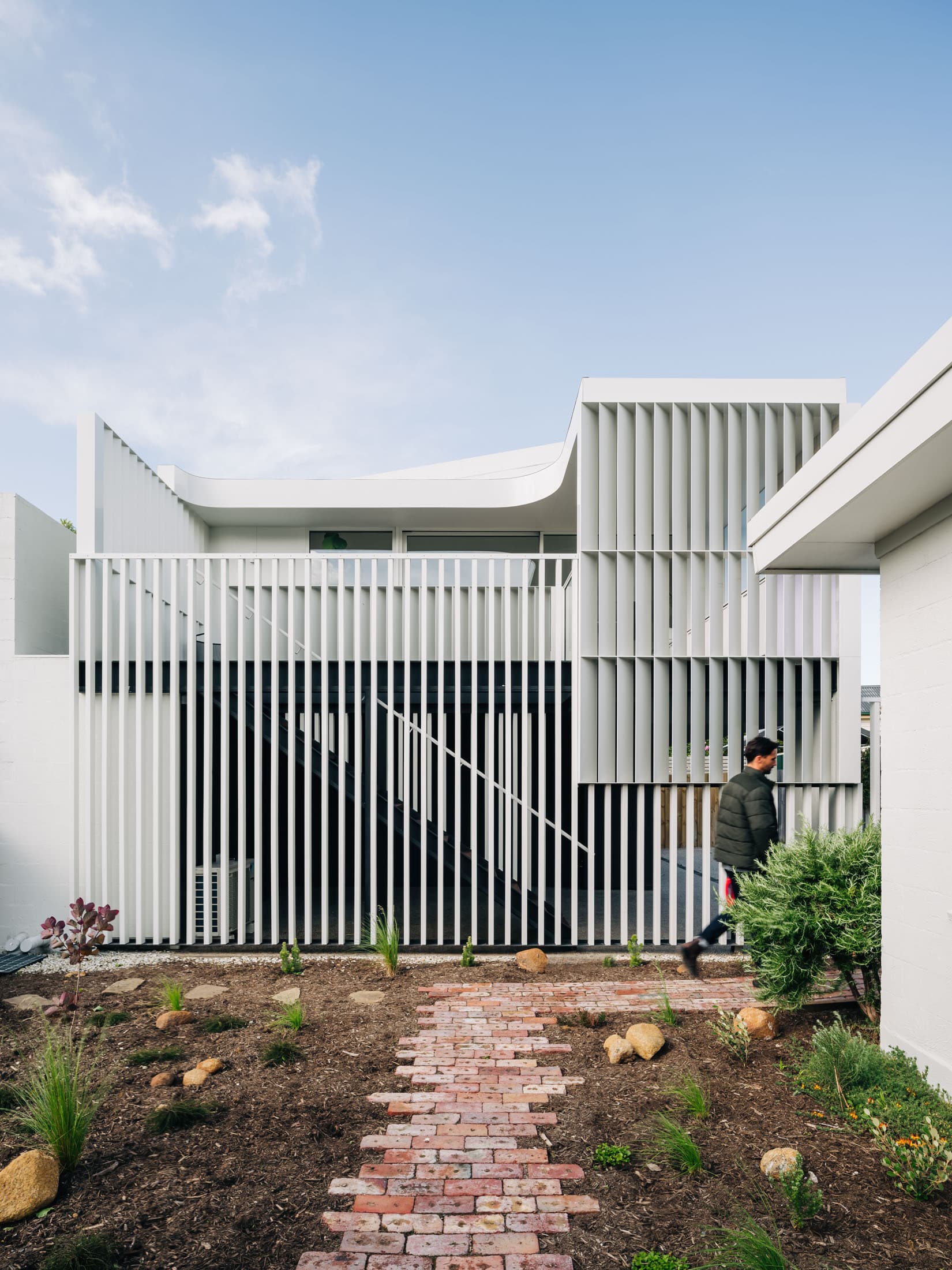 Man walks past a modern white house with vertical slats and a brick garden path.