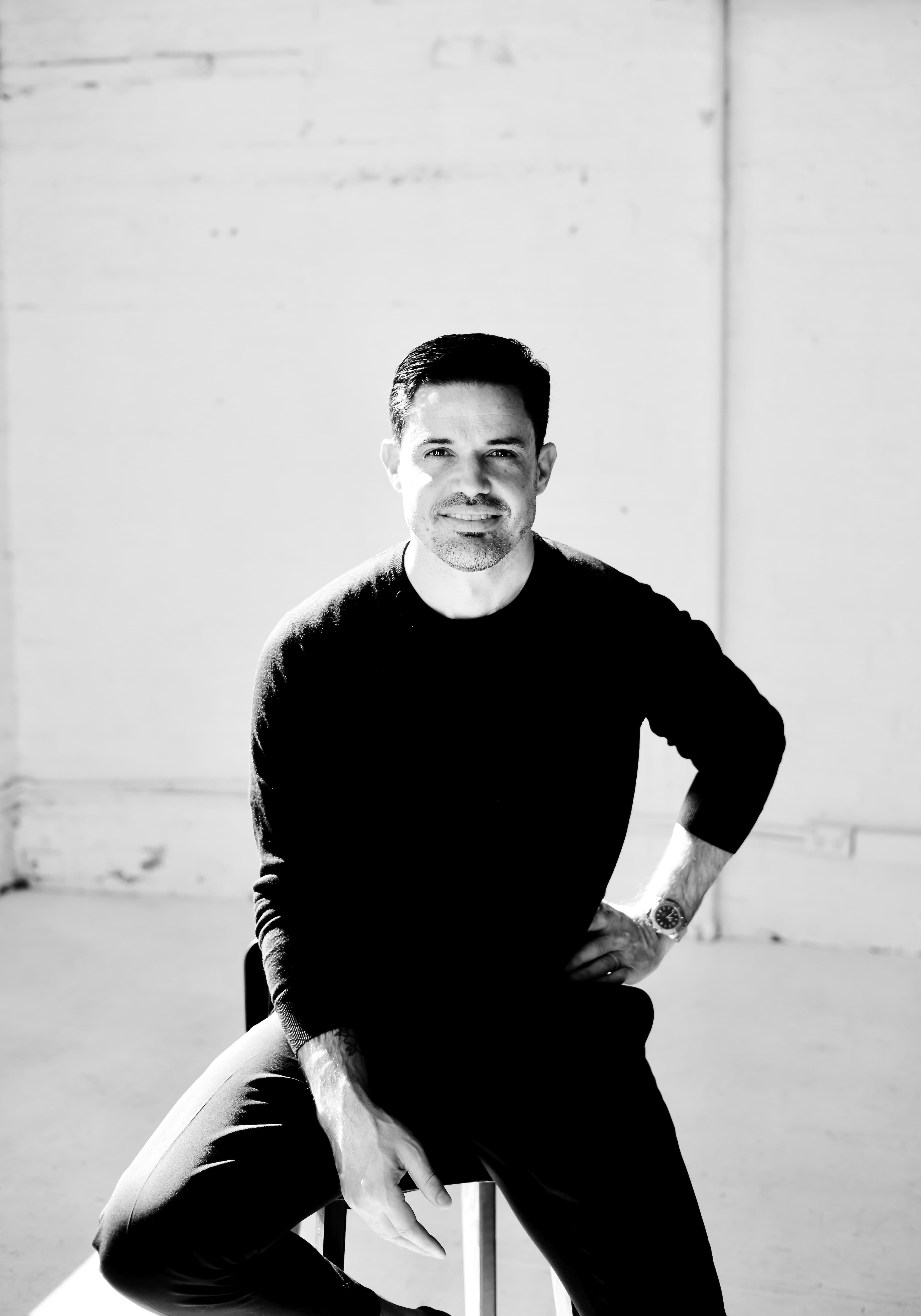 Black and white portrait of a smiling man sitting on a stool in bright light.