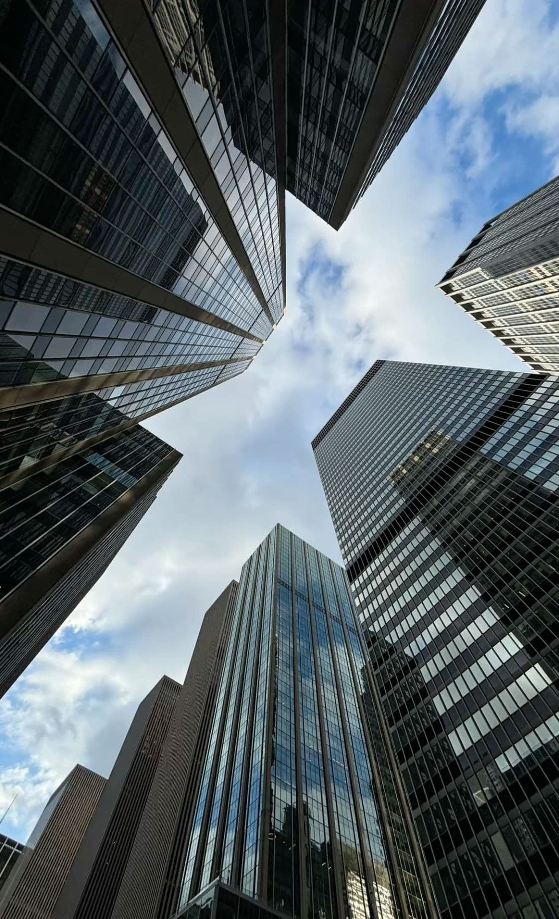 Low-angle view of towering glass skyscrapers reaching toward a blue sky with soft white clouds.