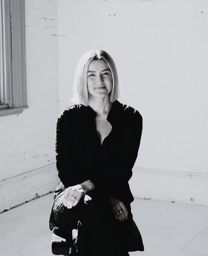 Smiling blonde woman in a dark shirt sitting in a bright white brick room.